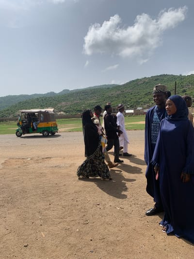 The image depicts a group of people standing on a dirt road in what appears to be Naraguta Hausawa, Nigeria. The terrain is arid, and there is a mountain in the background. A tuk-tuk sits parked on the road. The people are wearing a variety of clothing, including traditional attire.