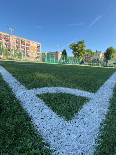 The image shows a close-up of a white corner marking on an artificial turf football field. The field is green and well-maintained. In the background, there are several apartment buildings, trees, and a fence enclosing the sports area. The sky is clear and blue with some wispy clouds. The scene is set in the city of Strzelce Opolskie, Poland, likely in a residential area or a public park, given the presence of apartment buildings. There are no people visible in the image.