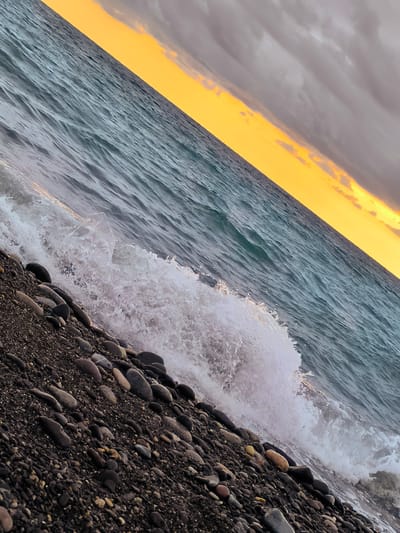 The image shows a rocky beach with waves crashing on the shore. The sky is filled with dramatic clouds, with a sliver of orange and yellow light from the setting sun peeking through. The water is a deep blue-green, and the pebbles on the beach are dark and varied in size. The scene is set at Zemo Kvariati, Georgia.