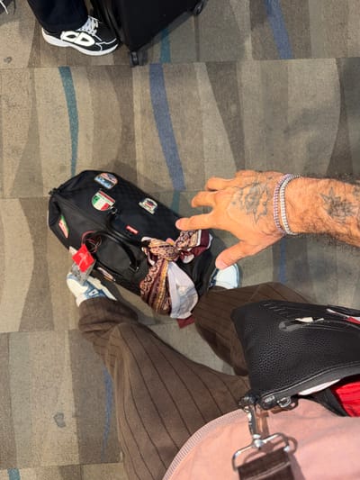 An overhead shot shows a person's legs and a black duffel bag on a patterned carpet. The person is wearing brown pinstriped pants and white and blue sneakers. A black handbag with a silver strap is resting on their lap. The duffel bag, which has travel stickers on it, is next to their left leg. The person's right hand, adorned with a pink and silver bracelet and tattoos, is visible reaching towards the duffel bag. The scene appears to be an indoor location, possibly an airport or hotel lobby, given the carpet and luggage. The location is not explicitly stated to be Mai Khao, Thailand.