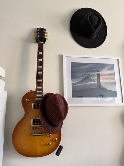 A Gibson Les Paul electric guitar is mounted vertically on a textured white wall, its sunburst finish facing the viewer. A brown plaid flat cap is draped over the body of the guitar, obscuring some of its lower portion. To the right of the guitar, a framed watercolor painting of a lighthouse is hung on the wall. The lighthouse is tall and slender, situated on a rocky outcrop with a mountain in the background shrouded in mist. The sky above the lighthouse is a dusky hue with hints of pink and orange. A black fedora-style hat is also mounted on the wall, positioned above and to the right of the framed painting. The environment appears to be an indoor setting, possibly a room in a home or studio. The image is taken from a medium distance, capturing the arrangement of these objects on the wall. The overall scene suggests a collection of personal items, perhaps reflecting the interests or style of the owner. The location is not specified in the image itself.