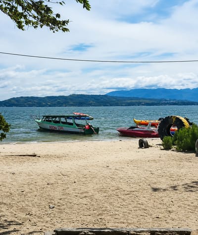The image depicts a serene lakefront scene in Lumban Bul Bul, Indonesia. In the foreground, a sandy beach stretches towards the water, with scattered driftwood. The calm lake is dotted with several boats, including a green and white passenger boat labeled "PESONA TUARA" and a smaller red boat with a yellow kayak on top. In the background, lush green hills rise against a backdrop of a cloudy blue sky. A tree branch with green leaves hangs from the top left corner of the frame, adding a touch of nature to the composition. Several large tires are visible on the shore, suggesting a recreational area.