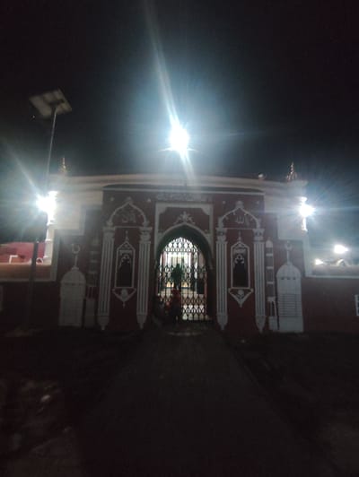 This is a nighttime, eye-level, outdoor, wide shot of a mosque entrance. The mosque is painted red with white decorative trim and Islamic calligraphy. An arched entryway is visible, covered by a black metal gate. Inside the gate, two figures can be seen. A solar-powered lamp is positioned to the left of the mosque, casting a bright light. The ground in front of the mosque appears to be dirt or gravel. The sky is dark. The mosque is located in Bagerhat, Bangladesh.