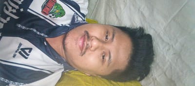 The image shows a close-up, sideways view of a young man lying on a light-colored surface, possibly a bed or sofa, with a yellow patterned cloth underneath. He is wearing a dark blue and white jersey with a graphic of a green crocodile head and the word "CROCODILE" in red letters. The jersey also has a logo with a stylized "M" and the text "OW BЯAA99A" in reverse. The man has dark hair, prominent eyebrows, and a mustache and stubble. His eyes are open, and his mouth is slightly parted, revealing his teeth. The background is a textured, light-colored fabric. The image's orientation suggests it might have been taken in Kemuning, Indonesia, where the subject is possibly located, but this cannot be definitively confirmed from the visual information alone.