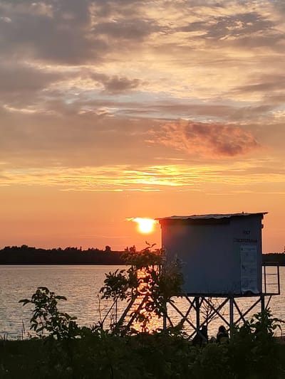 This is a medium-shot, eye-level, horizontal photograph of a sunset over a body of water, likely a lake or river, in Chelganovo, Russia. The sky is filled with dramatic, streaky clouds in hues of orange, pink, and grey. The sun, a bright white orb partially obscured by a thin cloud, is low on the horizon, casting a golden glow across the water. In the foreground, silhouetted against the bright sky and water, is a small, light blue lifeguard station raised on stilts. The station has a flat roof and appears to have some signage on its side, including Cyrillic text that reads "ПОСТ СПАСАТЕЛЬНЫЙ" (Lifeguard Post). There are also what appear to be informational posters on the side of the structure. Below the station, in the shadow of the structure and foreground foliage, two figures are seated or crouching. In the lower foreground, dark green foliage is visible, partially obscuring the view of the water and the base of the lifeguard station. The water surface reflects the colors of the sunset. The background horizon is a dark, silhouetted treeline. The overall mood of the image is serene and peaceful, capturing the end of a day in a rural setting.