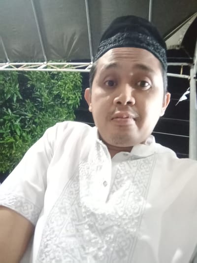 A medium shot selfie shows a man wearing a white tunic with decorative embroidery around the collar and down the chest. He is also wearing a black kufi on his head. The man has dark hair, brown eyes, and a light mustache. He is looking directly at the camera with his mouth slightly open. The background appears to be outdoors at night, with a dark sky, some greenery to the left, and a structure with a canvas roof overhead supported by poles. The overall setting suggests an outdoor gathering or event in Brangsong, Indonesia.