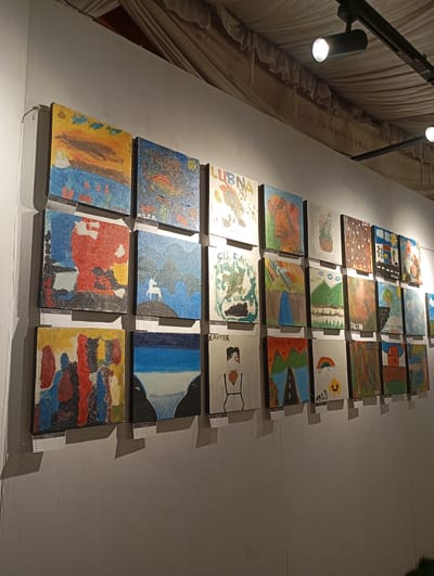 An art exhibition is displayed on a white wall in Meunasah Papeun, Indonesia. The artwork consists of numerous small, square paintings, mostly abstract or childlike in style, arranged in multiple rows. Some paintings have labels identifying them by name and artist. The lighting is provided by spotlights mounted on a track system above. The ceiling appears to be draped with fabric, creating a textured canopy. The overall scene depicts a display of local art in a gallery-like setting.