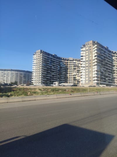 This media file captures a view of modern, high-rise apartment buildings under a clear blue sky. The buildings are characterized by their curved facades and numerous balconies, each with white railings. The architecture suggests a contemporary urban or suburban setting.

The foreground is dominated by a paved road with a grassy verge running alongside it. The road appears to be a street or an access road, with a concrete curb separating it from the grass. A dark shadow, possibly cast by a building or a large object, stretches across the road, indicating the presence of sunlight and therefore a daytime setting. There are hints of other structures or vehicles, such as a white truck and some parked cars, visible near the buildings. The presence of a faint contrail in the sky suggests air traffic overhead.

The scene appears to be a typical day in an urban or developed area. The clear blue sky and bright sunlight suggest fair weather. The architecture and the road indicate a planned development. The precise location is identified as Motodrom YS, Azerbaijan. No people are clearly visible. The overall impression is one of modern construction and a functional, residential environment. The image is taken from a slightly elevated perspective, looking upwards towards the buildings and the sky.