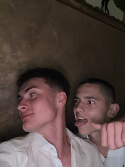 Two young males are captured in a dimly lit interior setting, with a dark, textured brown wall or ceiling forming the background. The lighting appears to be from a direct source, possibly a flash, illuminating the subjects’ faces and clothing against the dark environment.
The individual on the left, positioned in the foreground, is a young male with a light complexion and short, dark hair styled with a fade. He is seen in profile, looking to his left and slightly upwards, exhibiting a subtle, relaxed smile. He wears a white collared shirt, unbuttoned at the top.
The second individual, also a young male with a light complexion and very short dark hair, is positioned behind and to the right of the first, with his face partially obscured. He is looking towards the left, with his tongue extended in a playful gesture. His right hand is raised into the frame, with the index finger extended forward, appearing to point playfully towards the area near the first individual. He is also wearing a light-colored collared shirt, visible on his right side.
The background consists of an uneven, dark brown surface with subtle variations in tone and texture, suggestive of rough plaster or stone. In the extreme upper right corner, a faint, dark silhouette of two figures walking on a lighter path is barely discernible against a darker, possibly green-tinged, backdrop. This detail is highly obscured and does not provide clear environmental context.
The generic nature of the interior and background details in the image precludes any definitive visual confirmation of the specific location within the city of Slivnitsa, Bulgaria.