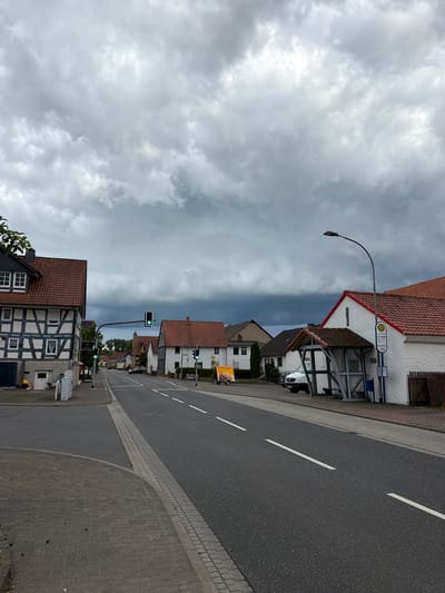 The image captures a street scene in Kirtorf, Germany, under a heavy, overcast sky. The street is paved with asphalt and marked with white lines. On the left side of the street, a traditional German half-timbered house with a red tile roof stands prominently, featuring dark wooden beams against white plaster. Adjacent to it, a sidewalk is paved with gray interlocking bricks. Across the street, a row of residential buildings with red tile roofs lines the road, interspersed with trees. A bus stop shelter with a gray metal frame and wooden benches is visible on the right side of the street, next to a white building. A street light with a modern design arches over the bus stop. Traffic lights are visible at an intersection, showing a green light. A few vehicles, including a white van and cars, are present on the street and parked. The overall atmosphere suggests an impending change in weather due to the dark, dramatic clouds.