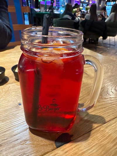 A red drink with a black straw in a handled mason jar is sitting on a wooden table. The glass is labeled "Le Burger" with a logo. In the background is a blurred view of a restaurant interior with people sitting at tables and bright lights. The location is Amstetten, Austria, specifically at a "Le Burger" restaurant.