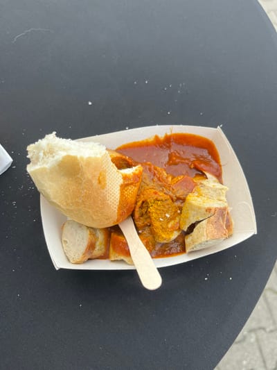 An overhead shot captures a classic German Currywurst served in a disposable white paper tray. The main dish consists of sliced sausage submerged in a rich, reddish-brown tomato-based curry sauce, heavily garnished with bright yellow curry powder and visible white specks, likely salt or additional seasoning. A small, light-colored wooden utensil, possibly a fork or spoon, rests partially in the sauce.
Accompanying the Currywurst are several pieces of crusty white bread, or a baguette/bun. A prominent piece of bread in the foreground shows a clear bite taken out of it, indicating it is being eaten. Other bread pieces are scattered within the tray, some covered in sauce.
The paper tray sits on a dark, possibly black, textured surface, which could be a table or another flat object. Small white crumbs or specks are visible on this surface, particularly in the lower left corner and around the tray, suggesting a casual eating environment. In the upper right corner and faintly along the top edge, a lighter, mottled surface, resembling pavement or a ground surface, can be seen, hinting at an outdoor setting. The lighting is bright and natural, suggesting daytime. The scene depicts a meal, likely enjoyed outdoors in Frechen, Germany, given the dish and context. There is no visible text.