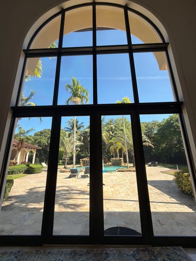 The image shows a view through a large, arched window with dark frames, looking out onto a sunny, tropical backyard. The window is divided into several panes. The upper part of the window reveals a bright blue sky with a few wispy clouds and the tops of palm trees.

The lower part of the window shows a paved patio area made of light-colored stones. To the left, there is a building with a covered walkway supported by columns, surrounded by manicured green hedges. Further into the yard, there is a swimming pool with a waterfall feature. Several lounge chairs are visible near the pool. There are numerous palm trees and lush green foliage throughout the yard, indicating a warm climate. The lighting suggests it is daytime, with strong shadows cast across the patio. The location is identified as Pinecrest, United States of America.