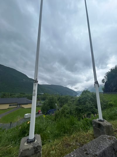 This image depicts a cloudy, overcast day in Røldal, Norway. The foreground features two white poles, likely part of a barrier or sign system, anchored to large grey stones. The poles are positioned diagonally across the frame, with the lower pole extending towards the right and the upper pole extending towards the left. The ground adjacent to the poles is covered in lush green grass, with some patches of brown earth and scattered wildflowers.

In the midground, a village or cluster of buildings is visible, nestled against a backdrop of steep, verdant mountains. The architecture appears to be a mix of traditional and modern styles, with a prominent yellow building with dark roofing and a smaller, darker building with white-framed windows. A red vehicle is parked on a road leading through the village. Trees are abundant, covering the lower slopes of the mountains and lining the edges of the village.

The background is dominated by dramatic, cloud-covered mountains, hinting at the rugged and majestic natural landscape of Norway. The sky is a uniform grey, suggesting a cool, possibly damp, and overcast weather condition, typical for this region. There is no visible text in the image. The overall impression is one of a serene, natural environment under a somber sky, with human presence evident in the form of buildings and infrastructure.