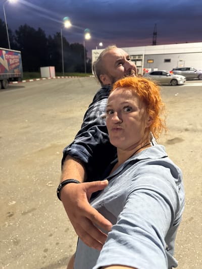 A man and a woman are taking a selfie outdoors. The woman, with vibrant red, curly hair, is in the foreground, making a kissy face at the camera. She is wearing a light blue long-sleeved shirt. The man, with short, graying hair and a beard, stands behind her to her left. He is wearing a dark and light plaid long-sleeved shirt. His left arm is visible, and he is wearing a dark braided bracelet on his wrist. The background suggests an evening setting. A building with metallic siding is visible on the left, with a few cars parked in front of it. Streetlights illuminate the scene. Further back, trees are visible against a dusky sky. A truck with colorful graphics on its side is parked in the lower right portion of the image, and a red and white striped border runs along the edge of the pavement. The image appears to be taken in Можга, Russia. The text on the truck is partially visible but not legible.