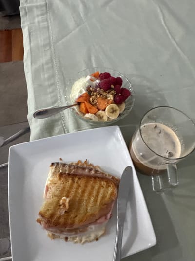 An overhead image depicts a meal set on a light green tablecloth in Nogueira e Silva Escura, Portugal. A white rectangular plate presents a toasted sandwich, appearing to contain ham and melted cheese, with a single bite removed from its left side. A silver butter knife rests to the right of the sandwich on the plate. To the upper right, a clear glass bowl holds a fruit and yogurt parfait composed of a white base (likely yogurt or cream), topped with red raspberries, sliced orange-colored fruit (possibly apricot or persimmon), sliced bananas, and granola or seeds. A silver spoon is positioned within the fruit bowl. To the lower right, a clear glass mug with a handle contains a coffee beverage topped with a layer of foam. The surrounding environment beyond the tablecloth is a softly blurred indoor area with muted tones. No individuals are visible.