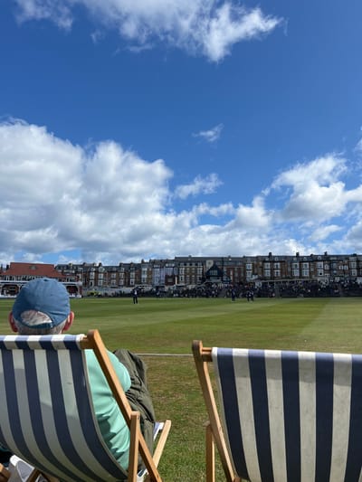 The image depicts a cricket match in progress at the North Marine Road Cricket Ground in Scarborough, United Kingdom. In the immediate foreground, from a spectator's perspective, the back of an individual seated in a blue and white striped wooden deck chair is visible. This person wears a light blue baseball cap and a light green shirt, with a dark jacket draped behind them. The back of a second, empty, identical deck chair is positioned to the right. A white boundary rope separates the foreground from the well-maintained green cricket pitch, where several players are active. Beyond the pitch, a significant crowd of spectators is gathered in tiered seating and on a grassy embankment. A long row of multi-story, traditional brick and stone terrace buildings, consistent with the architecture along North Marine Road, forms the backdrop to the cricket ground. To the far left, a building with a distinctive red-tiled roof, likely the cricket pavilion, is partially visible. The sky above is bright blue with scattered white cumulus clouds.