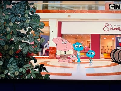 The image shows an animated scene from a cartoon set in a shopping mall. In the foreground on the left is a large, leafy green plant. To the right, four cartoon characters are standing on a polished floor with curved orange and white lines. The characters include a large pink rabbit-like figure, a smaller pink figure, a blue cat-like figure wearing a shirt and shorts, and another blue cat-like figure with a bowed head. Behind them is the facade of a store with the word "Shoe" in stylized red letters. Above the store, a Cartoon Network logo is visible. The background shows the interior of a mall with marble accents and glass windows displaying merchandise. A man in a suit is seated on a stool in the background, to the left of the frame. The location is not Topolovgrad, Bulgaria.