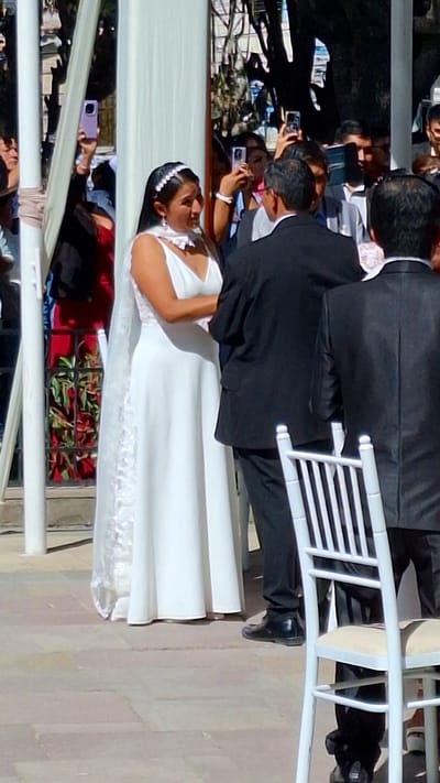A wedding ceremony is in progress in Cerro Colorado, Peru. The bride, in a white dress and a beaded headpiece, stands embraced by the groom. He is in a black suit. Guests are taking pictures with their phones. A row of floral arrangements is visible. White chairs line the event space.