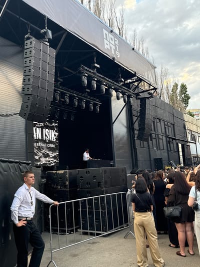 The image depicts an outdoor event stage, likely in the city of Kozelsk, Russia, given the context. In the foreground, a security guard in a white shirt and black pants stands near a metal barrier, looking to the left. Behind him, a crowd of people, mostly women, are gathered, facing the stage. The stage itself is large and black, with rows of speakers stacked on either side. A banner displaying "AN ISIK INTERNATIONAL" hangs prominently on the left side of the stage. A DJ is visible on the stage, operating equipment. Above the stage, a large black structure with the logo "NO2 PT.9" is mounted. The background shows bare trees and a cloudy sky, suggesting an autumn or early spring setting. The overall scene suggests an outdoor concert or festival.