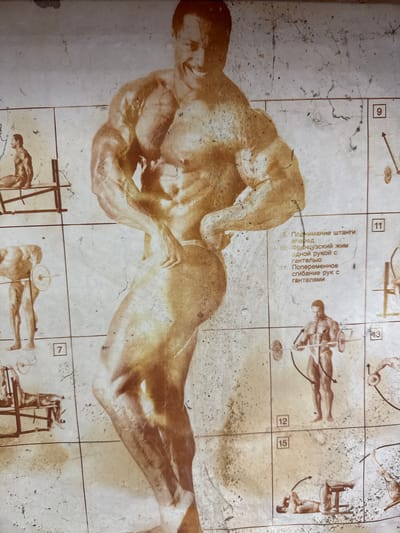 The image is a faded, sepia-toned poster depicting a bodybuilder and various weightlifting exercises. The central figure is a large, muscular man in a flexing pose. Smaller illustrations of exercises are arranged in a grid around him, each with a number and text in Cyrillic. The poster appears to be old and has cracks and stains, suggesting wear and tear. The location is identified as Khazar Raion, Azerbaijan, which is a district of Baku, the capital city. The poster itself is the subject, a visual guide to bodybuilding.