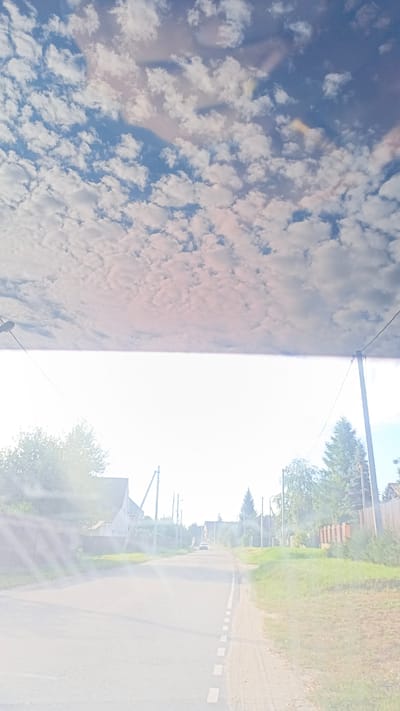 The image is a vertical, slightly tilted, first-person perspective shot looking down a paved rural road in the city of Zuy, Russia. The upper half of the image features a bright blue sky filled with scattered white cumulus clouds, with some pinkish hues suggesting early morning or late afternoon light. The lower half of the image shows the road stretching into the distance, flanked by green grass, trees, and wooden fences. Several utility poles with wires are visible along the sides of the road. In the distance, a white car is approaching on the road. Houses with pitched roofs are visible behind the fences on the left side of the road. The scene appears to be well-lit, with some lens flare effects creating streaks of light across the lower left portion of the image.