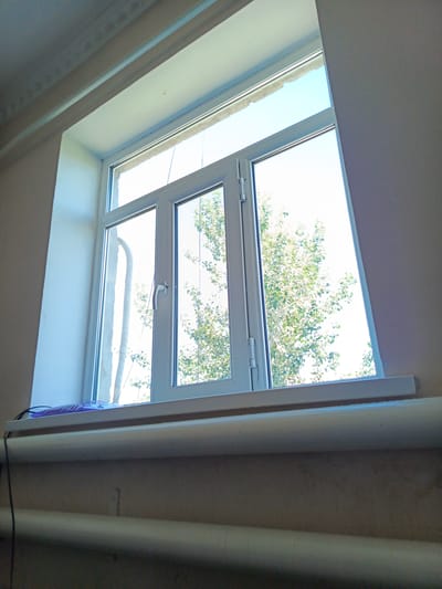 A low-angle interior shot captures a modern white uPVC casement window with three panels. The leftmost panel is a fixed pane, while the middle and right panels are operable, featuring white handles. Outside the window, lush green foliage of a tree is visible against a bright blue sky. The interior features a white windowsill with a purple object and black cables resting on it. Below the windowsill, two horizontal white pipes, likely radiators, are prominent against a beige wall. The upper part of the window frame is framed by a light-colored, textured ceiling molding. The overall impression is of a room in a building, potentially an apartment or an office, in Nukus, Uzbekistan, with a view of natural greenery.
