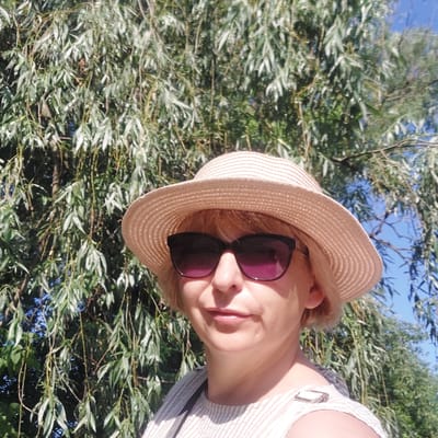 A medium shot captures a woman wearing a straw hat and sunglasses, facing the camera with a slight smile. She is outdoors, with a backdrop of lush green foliage, likely willow trees given their drooping branches. Sunlight filters through the leaves, creating dappled light on her face and clothing. The woman is wearing a striped top. The sky is visible in patches through the branches, suggesting a bright, clear day. The location is identified as Voronkiv, Ukraine.