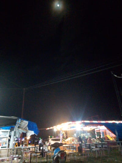 The image depicts a nighttime scene at what appears to be a fair or market in Gampongpaya, Indonesia. The sky is dark, with a prominent, bright moon visible in the upper left quadrant. Below the sky, there are several structures and objects.
On the left, there is a stall or booth with a blue tarp covering a portion of it, and shelves visible inside. To the right of this, there are a series of tables and chairs, suggesting a seating area. Beyond these, a brightly lit structure is visible, possibly an amusement ride or another vendor stall, adorned with colorful lights. There are several people present, some standing and some possibly seated at the tables. One person in the foreground appears to be standing near a motorbike. The overall impression is one of a lively evening gathering.
The scene is illuminated by artificial lights from the stalls and rides, creating a contrast with the dark night sky and the bright moon. There are also power lines visible stretching across the mid-ground. The setting suggests an outdoor event taking place after dark. No visible text is discernible in the image.