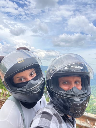 In the city of Son Dien, Vietnam, two individuals are captured in a close-up, selfie-style photograph while wearing motorcycle helmets. The person on the left, positioned slightly closer to the camera, wears a black helmet adorned with a gold crown emblem and the word "Royal" printed in gold lettering. Their eyes are visible above a black face mask, and their brown hair is styled with two pom-pom-like accessories. They are wearing a white top with a light green strap across their chest.
The person on the right wears a black helmet with a clear visor, displaying the "LS2" logo. Their blue eyes are visible, and they are wearing a black nose guard. They are dressed in a grey and black plaid shirt. Both individuals appear to be looking towards the camera.
The background of the image reveals a lush, green landscape with rolling hills and trees, indicative of a rural or mountainous area. The sky above is filled with scattered white clouds against a bright blue backdrop, suggesting daylight conditions. A blurred effect on the background hints at the subjects being in motion or the focus being intentionally on the individuals.
