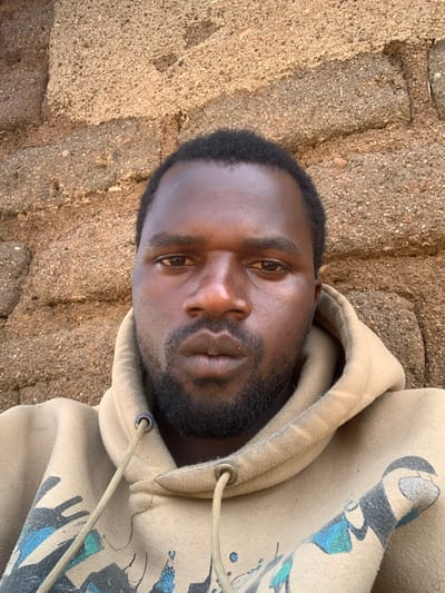 The image is a head-and-shoulders selfie of a Black man with dark skin and short, black hair, and a beard. He is wearing a light brown hooded sweatshirt with drawstrings. The background consists of a rough, textured wall made of brown bricks or mud. The man's face is centered in the frame, looking directly at the camera. The lighting appears natural and somewhat harsh, casting subtle shadows on his face. The setting is an outdoor environment. Based on the provided information, this scene is located in Buga da Gindi, Nigeria.