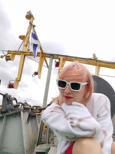The image shows a woman with pink hair, wearing white sunglasses and a white sweater with a beige pattern. She is sitting on the deck of a boat, with her chin resting on her hand. In the background, there is a yellow structure with a blue and white flag. The sky is overcast, suggesting a cloudy day. The woman appears to be pensive. The visible text is "Novokemskii, Russia", which is the location context provided.