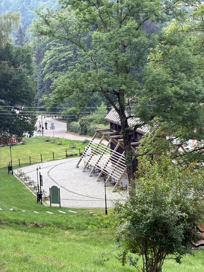 The image captures a scene in Świeradów-Zdrój, Poland, focusing on a park-like area with a prominent wooden structure. In the foreground, a neatly paved circular area is bordered by a black metal fence and several park benches. A large tree stands to the right, partially obscuring a unique wooden building constructed with inclined beams and a roof. This structure appears to be part of a salt graduation tower, a common feature in Polish spa towns, designed to release aerosolized salt water for therapeutic purposes. Beyond the paved area, a grassy slope leads up to a road where two individuals are visible, one standing on a self-balancing scooter and another walking. Several more benches are scattered along the road, with trees and lush greenery forming the backdrop, suggesting a natural, possibly mountainous, setting characteristic of the Karkonosze region. The overall atmosphere is serene, with a muted color palette hinting at an overcast day.