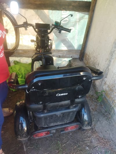 The image shows a black three-wheeled electric scooter, possibly a mobility scooter, parked outdoors. The scooter is mostly black with some chrome accents. It has a large seat, handlebars with controls, a digital display, and a basket at the front. The rear of the scooter features red taillights and a reflective strip. The scooter appears to be somewhat worn, with scuffs and scratches visible on its body.
To the left of the scooter, a portion of a bicycle wheel and a person wearing a red t-shirt and blue pants can be seen. The person is also wearing a beige slipper on their right foot. A bright green tarpaulin or fabric is draped over something behind the scooter. The scooter is parked on a dirt and grass surface, with a concrete or stucco wall on the right side of the frame. Dry grass and some small green plants are also visible around the scooter.
In the background, there is a wooden structure and a large sheet of what appears to be plastic sheeting or tarp with some text printed on it. The text on the sheeting is partially visible and seems to be in Cyrillic characters. Part of the text reads "NICE ON HAШIA" and below that "BHKEDIM".
The lighting suggests it is daytime, possibly late morning or early afternoon, with sunlight casting shadows. The scene appears to be an outdoor, possibly rural or semi-rural, setting in Sosnivka, Ukraine. There is no specific activity taking place, and the scooter appears to be stationary. The overall impression is one of a functional, albeit used, mobility device parked in a utilitarian outdoor space.