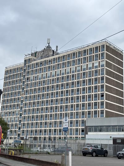 The image shows a large, multi-story building with a facade of concrete and numerous windows. The building has a flat roof with several antennas and communication equipment. At the base of the building, there is a sign that reads "Rail House." The surrounding area appears to be a parking lot with several cars visible, including a dark grey hatchback and a silver Audi. A metal fence encloses the parking area. The sky is overcast. The building is located in Crewe, United Kingdom.