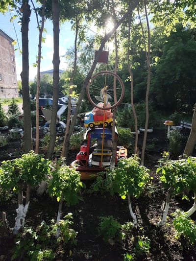The image depicts an outdoor scene within a city, possibly Petropavl City Administration, Kazakhstan, featuring a garden art installation made of recycled materials. The installation is centrally located and appears to be a stacked arrangement of various objects, including toy trucks, what seems to be a red tractor-like object, and possibly other discarded items like tires, all painted in bright colors. A bicycle wheel is suspended above the installation. In the background, a multi-story building with visible windows is present, suggesting an urban environment. The foreground is dominated by lush green foliage, with several trees featuring white-painted lower trunks, indicating a garden or park setting. The scene is illuminated by sunlight filtering through the trees. The overall impression is one of a whimsical, self-made art display in a residential or communal outdoor space.