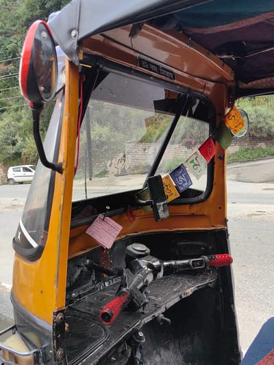 The image shows the interior and side of a yellow auto-rickshaw parked on a road. The auto-rickshaw has a black canopy. A red and black rearview mirror is visible on the left side. Prayer flags with Tibetan script hang across the windshield. A pink slip of paper, possibly a receipt or permit, is pinned to the dashboard. The steering mechanism and handlebars are visible, with a red grip on the right handlebar. The exterior of the auto-rickshaw is yellow and black. In the background, a white car is parked on the side of the road, and a brick wall and lush greenery are visible. The overall scene appears to be in a rural or semi-urban environment. The location is identified as Siyal, India.