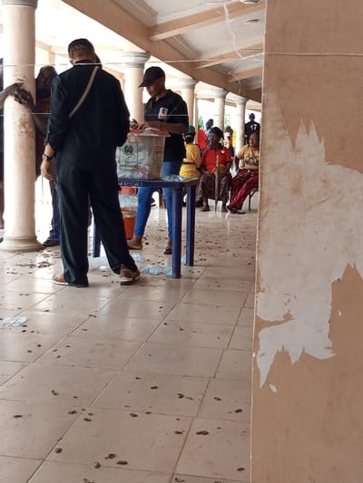 The scene depicts an outdoor or semi-enclosed public area, possibly a veranda or covered walkway, identified as being in the city of Ajalli, Nigeria.
In the foreground, a light brown wall on the right exhibits significant paint peeling, revealing lighter patches underneath. The tiled floor is light beige with a grid pattern, covered with numerous small, dark organic fragments resembling peanut shells and several crumpled clear plastic water sachets.
In the mid-ground, a dark blue plastic table serves as a focal point. On the table rests a transparent plastic ballot box, partially filled with papers. The ballot box features a green logo, identifiable as the Independent National Electoral Commission (INEC) symbol of Nigeria, and includes a slot for inserting ballots. Below the table, a white plastic bag or container displays the text "MAMI RENTALS." Two individuals are positioned around this table. One individual, seen from the back, wears a dark long-sleeved shirt, dark trousers, and light-colored shoes, with a light strap across their back. They are facing towards the table, interacting with its contents. The second individual, to the right of the first, wears a black t-shirt, a black baseball cap, and blue jeans, standing behind the table and appearing to organize items on its surface.
In the background, a series of light-colored, round architectural pillars with decorative caps support an overhead structure. Several people are visible further back. To the far left, a person leans against a pillar. To the right, three individuals are seated: one older woman in a red top and patterned skirt holding a walking stick or umbrella, another woman partially obscured in a yellow garment, and a third person in a dark t-shirt with the number "17" printed on the back. Additional blurred figures are visible in the distant background. The overall lighting is bright and even, indicating an outdoor or well-lit environment.