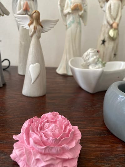 The image displays a collection of angel figurines and a pink rose figurine on a dark wooden surface. In the foreground, a large, detailed pink rose figurine is centrally placed. Behind the rose, several angel figurines are visible, varying in size and design. One prominent angel figurine in the middle ground is light-colored with outstretched wings and hands clasped in prayer, featuring a white heart emblem on its torso. To its left, another tall, light-colored angel figurine with hands in prayer is partially visible. Further back, other angel figurines are discernible, with one in the far background appearing more ornate. A small, white angel figurine is situated in a white, heart-shaped dish. To the right of the rose, a portion of a smooth, grey, cylindrical object is visible, possibly a vase or pot. The background is out of focus, showing a light-colored wall. This scene is not located in the city of Osek, Czechia.