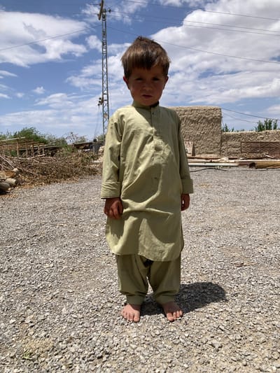 A barefoot boy in a light green traditional shalwar kameez stands on a gravel path in Qilla Saifullah, Pakistan. The sky is visible with clouds, and there is a structure made of mud or thatch in the background. There are power lines visible and stacked branches.