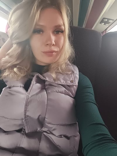 A young woman with long, wavy blonde hair and a direct gaze takes a selfie inside what appears to be a public transport vehicle, situated in the city of Sovkhoza "Pyatiletka", Russia. She is wearing a dark green, ribbed long-sleeved top underneath a light lavender or pale purple quilted puffer vest, which is partially zipped. Her left hand is raised, with her fingers touching her hair near her temple. In the blurred background on the left, the top of another person's head is partially visible. The interior of the vehicle features dark, possibly black, upholstered seats and an overhead ceiling panel system with distinct green, gray, and purple-toned stripes, along with a white rectangular overhead light or ventilation unit containing four circular elements. Soft, bright light enters from the upper left, suggesting a window.