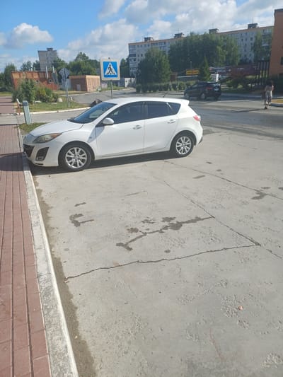 A white hatchback is parked on a cracked concrete surface next to a brick sidewalk. In the background, several multi-story apartment buildings and a commercial building with a "Пятёрочка" sign are visible. A pedestrian crossing sign is located to the left of the frame. A dark-colored SUV is driving away from the camera in the distance, and a woman in a light-colored dress is walking in the same direction. The sky is blue with scattered clouds. The scene appears to be in an urban environment within Elitnyi, Russia, based on the architecture and signage.