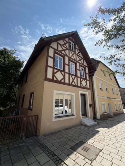 In Ebermannstadt, Germany, on a sunny day with scattered clouds, a traditional half-timbered building stands prominently on the left, its facade a mix of tan plaster and dark brown timber framing with diamond-shaped infills. The building features several windows, some with white frames. To its right, a yellow building with white trim runs parallel, also displaying multiple windows with white roller shutters. A paved walkway, made of grey rectangular stones and bordered by smaller dark stones, runs in front of both buildings. A rusty metal fence and dense green foliage are visible to the far left. Sunlight casts strong shadows across the walkway, illuminating a metal grate in the foreground.