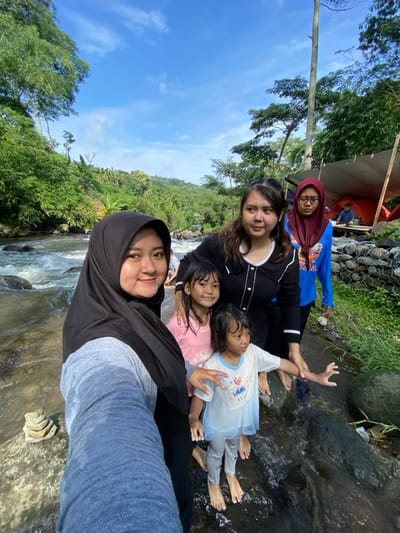 A group of five women and children pose for a selfie along a rocky stream. The woman in the foreground wears a black hijab and grey top. Behind her, three women and two young children are partially submerged in the water. The backdrop features lush green trees and a bright blue sky. In the upper left corner, a glimpse of camping tents is visible. The photograph is taken from a low angle, emphasizing the height and depth of the natural setting.