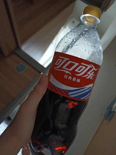 A person's hand holds a clear plastic bottle of Coca-Cola, condensation visible on the bottle's surface. The bottle is predominantly filled with a dark liquid and has a red label with white Chinese characters, which translate to "Coca-Cola Classic Taste." The background is blurred, showing wooden furniture and a light-colored wall, suggesting an indoor domestic setting. The image appears to be taken in Hongshan District, China.