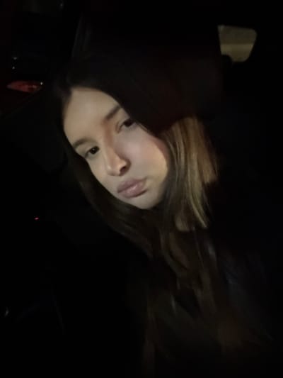 A woman with light skin and long, wavy hair, partially highlighted blonde, is visible. Her face is angled slightly towards the viewer, with dark eyes and full lips. The background is predominantly dark, with indistinct shapes suggesting an interior space or a vehicle. A section of dark fabric, possibly clothing, is also visible. The lighting is low, casting shadows across the scene.