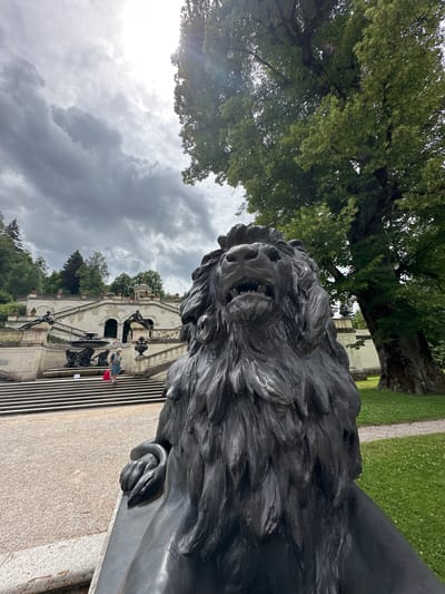 The image captures a view of the Linderhof Palace gardens in Ettal, Germany. In the foreground, a dark bronze lion sculpture is positioned prominently, its detailed mane and fierce expression clearly visible. To the left and in the mid-ground, a grand staircase leads up to a white, multi-tiered building adorned with statues and urns. A few people are present on the stairs, one individual carrying a red bag. The background features lush green trees and a cloudy sky with sunlight breaking through. The overall scene depicts an outdoor setting with classical architecture and natural elements.