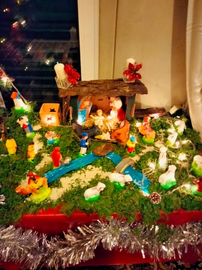 A vibrant Nativity scene unfolds on a red-covered surface adorned with silver tinsel. A wooden stable shelters Mary, Joseph, and baby Jesus. Various painted figures representing shepherds, animals, and other characters are scattered across a green mossy landscape. A blue plastic river meanders through the scene, and small white lights are dispersed among the figures and moss. A lit star lantern adds to the warm glow, and candles with red accents adorn the top of the stable.