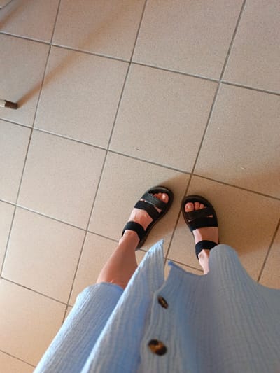 The image shows a person's legs and feet, wearing black sandals and a light blue textured garment. The feet are standing on a tiled floor with light beige square tiles and gray grout lines. The perspective is from above, looking down. The lighting suggests an indoor environment. There are no discernible elements that would indicate a specific location within the city of Zalesovo, Russia.