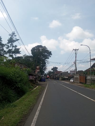 The scene depicts a straight asphalt road extending into the distance under a blue sky with scattered white clouds, located in Niz Lakuk, India. The road features a solid white shoulder line on the left and a dashed white center line. On the left side of the road, an elevated grassy bank with dense green foliage runs alongside a concrete ditch. Beyond the bank, multiple structures, including small shops or residences, are visible. One prominent building displays an aged mural and multiple signs, one of which indicates "DISCOK LAT M NATA". A blue van is parked near these structures. Tall pine-like trees and other broadleaf trees are present on the left, with numerous utility lines strung overhead. On the right side of the road, a white compact car is parked on the shoulder, followed by a concrete fence with a brown wooden gate. A utility pole heavily laden with wires stands on the right, alongside a modern streetlight. In the distant midground, indistinct figures, possibly two individuals on bicycles or motorcycles, are observed moving away from the camera. Additional residential buildings line both sides of the road in the background.